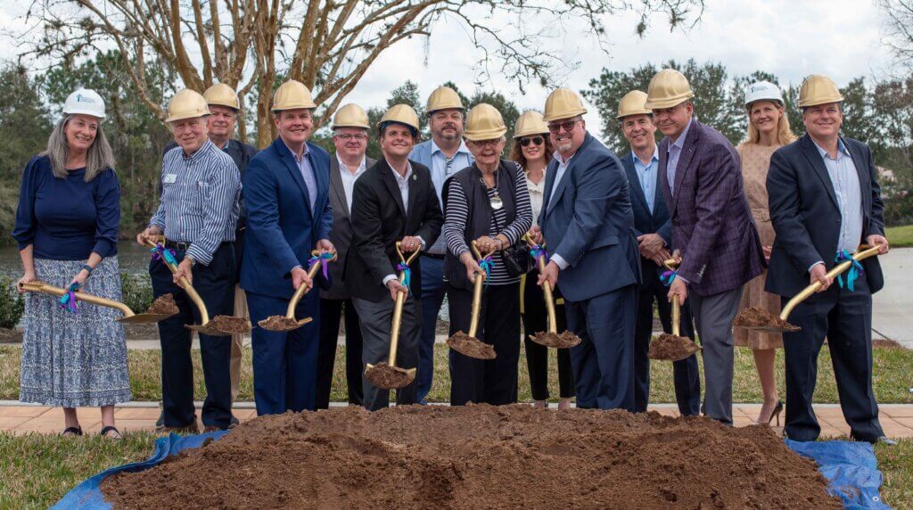 Several people hold loaded shovels above dirt pile for groundbreaking ceremony